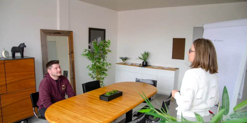Ein an einem runden Holztisch sitzenden Coachingteilnehmer spricht mit dem stehenden Coach